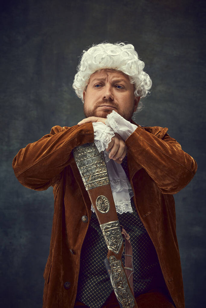 Sad. Portrait of young man in image of nobleman in vintage brown hunting suit and white wig with old hunting rifle isolated over dark retro background. Art, fashion, emotions, male hobby concept - Фото, зображення