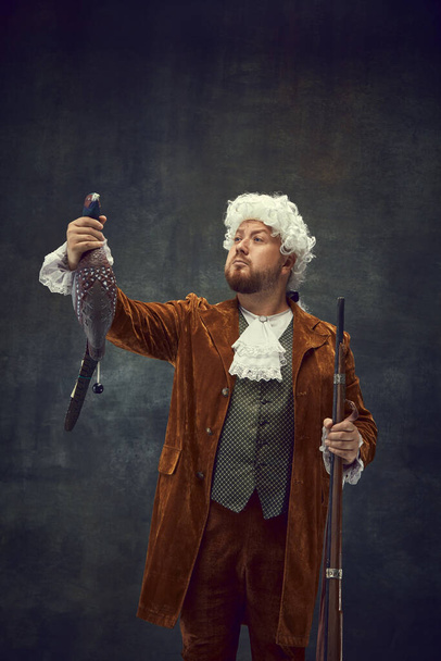 Hunter with funny prey. Young man in image of nobleman in vintage brown hunting suit and white wig with old hunting rifle isolated over dark retro background. Male hobby, humor concept - Fotografie, Obrázek