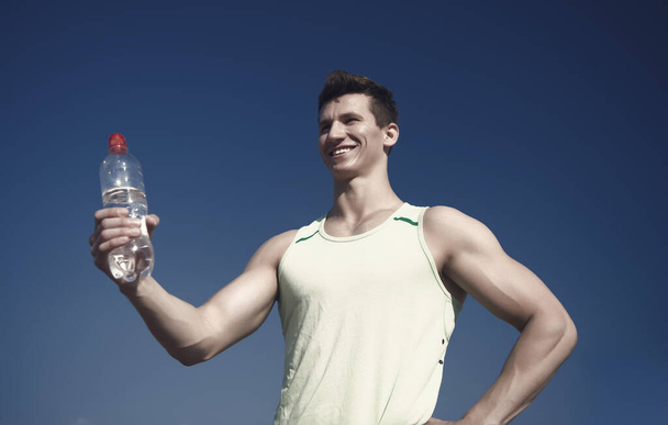 Happy man with water bottle in muscular hands. Bodybuilder with muscles biceps, triceps on sunny day. Healthy lifestyle and health. Sport and fitness concept. Athlete in green tshirt on blue sky. - Fotó, kép
