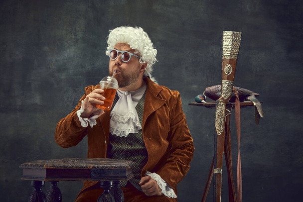 Cocktail party. Vintage portrait of young man in brown vintage suit and white wig like medieval royal hunter isolated on dark background. Comparison of eras concept, renaissance style. - Foto, Bild