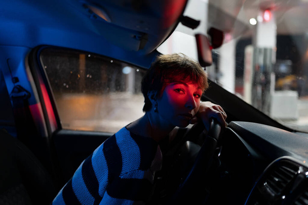Tired Caucasian woman sits behind wheel with head resting on steering waiting for fellow traveler. Girl driver with neon red light on face is dressed in striped sweater in line for refueling car - Фото, изображение