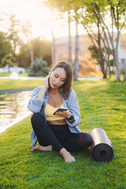 Slim mulher desportiva segurando telefone celular em sua mão enquanto sentado no parque da cidade perto do tapete de exercícios. Comunicação por telemóvel. Treino de fitness. Aptidão - Foto, Imagem