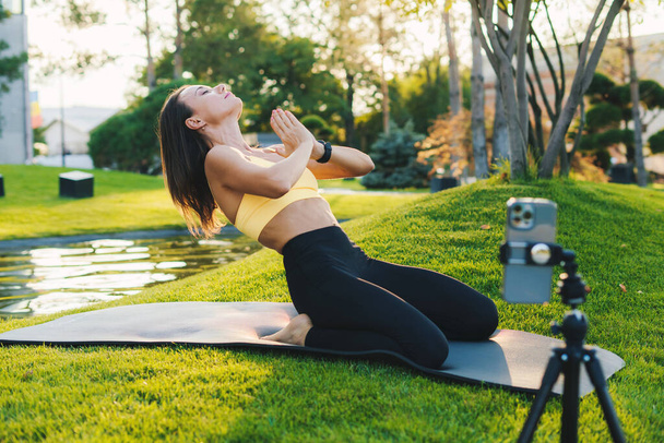 Treinador de fitness atlético em sportswear filmagem de vídeo no telefone da câmera como ela fazendo ioga no tapete no parque de verão ao ar livre. Estilo de vida saudável, desporto. Aptidão - Foto, Imagem