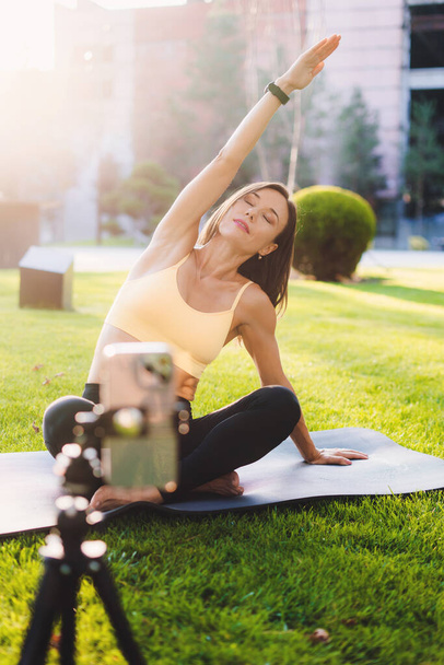 Young woman blogger in a sport wears using webcam recording video for her blog practicing yoga in the morning in the park. Fitness blog. On the grass in city - Foto, Imagen