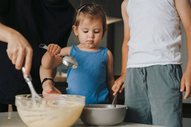 Matka uczy małe urocze dzieci mieszania mąki z mlekiem, gotowania razem w nowoczesnej kuchni. Proces gotowania. Szczęśliwa rodzina, dzieciństwo. - Zdjęcie, obraz