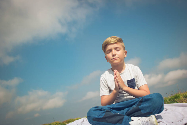 Small boy in namaste position meditating with eyes closed on a meadow against the sky. Copy space.  - Photo, Image