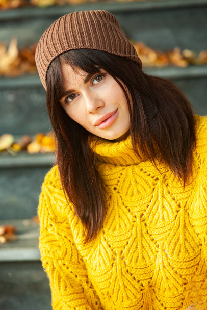 Beautiful European brunette dressed in a yellow woolen sweater and hat outside. The beautiful girl who wears thick stylish autumn clothes in cool weather. - Fotoğraf, Görsel