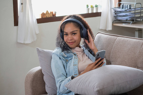 An African American woman uses a smartphone and puts on a music headset in her home to relax. - Foto, Imagen