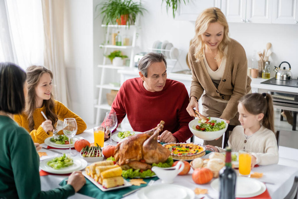 Smiling woman holding salad near daughter and family during thanksgiving dinner  - Φωτογραφία, εικόνα