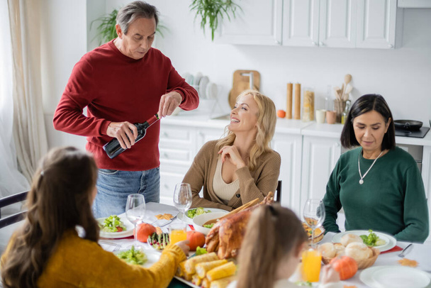 Улыбающаяся женщина смотрит на родителя с вином рядом День благодарения ужин и мультиэтническая семья дома  - Фото, изображение