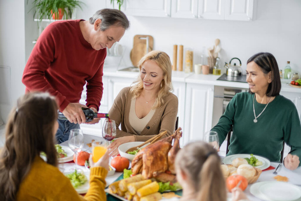 Uomo anziano versando vino vicino sorridente famiglia multiculturale durante la cena del Ringraziamento a casa  - Foto, immagini