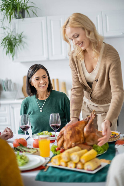 Multicultural mother sitting near daughter putting festive turkey on table at home  - Photo, Image