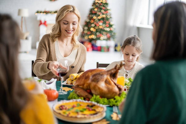 Smiling woman holding glass of wine near family and blurred thanksgiving dinner at home  - Photo, Image