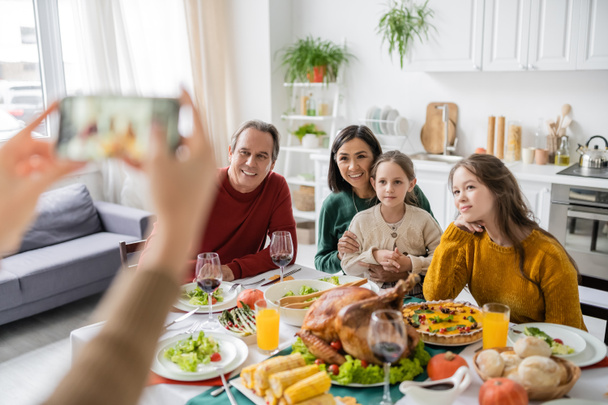 Multiethnic family sitting near thanksgiving dinner while woman taking photo on smartphone at home  - Photo, Image