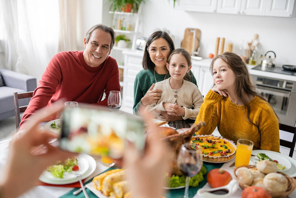 Interracial family smiling near delicious thanksgiving dinner and woman taking photo on smartphone at home  - Foto, Imagen