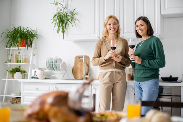 Smiling multiethnic mother and daughter holding glasses of wine during thanksgiving celebration at home  - Photo, Image