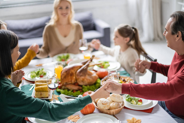 Interracial grandparents holding hands near blurred family and thanksgiving dinner at home  - Photo, Image