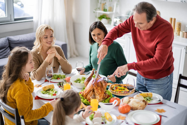 Man cutting turkey near smiling multiethnic family during thanksgiving dinner at home  - Photo, Image
