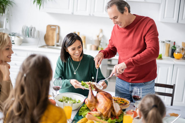 Cheerful grandparent putting turkey on plate near multicultural women and kids during thanksgiving dinner  - Photo, Image
