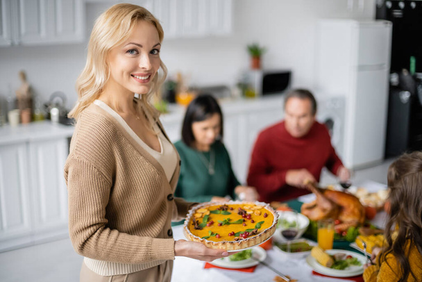 Smiling woman holding thanksgiving pie and looking at camera near blurred family at home - Foto, Imagen