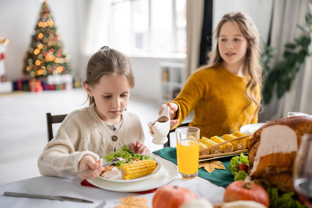 Κορίτσι κοιτάζοντας σαλάτα και καλαμπόκι κοντά θολή αδελφή με σάλτσα κατά τη διάρκεια των Ευχαριστιών δείπνο στο σπίτι  - Φωτογραφία, εικόνα
