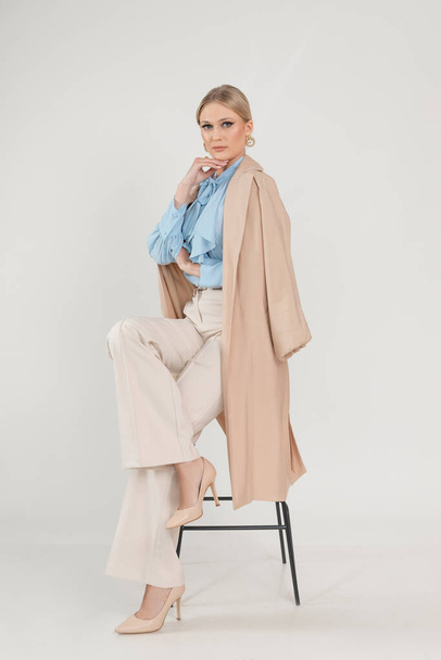 Full length photo of beautiful young woman sitting on chair in delicate cloak and pants, white background, free copy space. Business people, basic female clothes, casual office outfit, seasonal look. - Fotografie, Obrázek