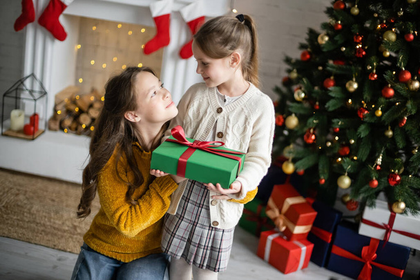 happy sisters holding gift box and looking at each other near blurred fireplace and christmas tree in living room - Photo, image