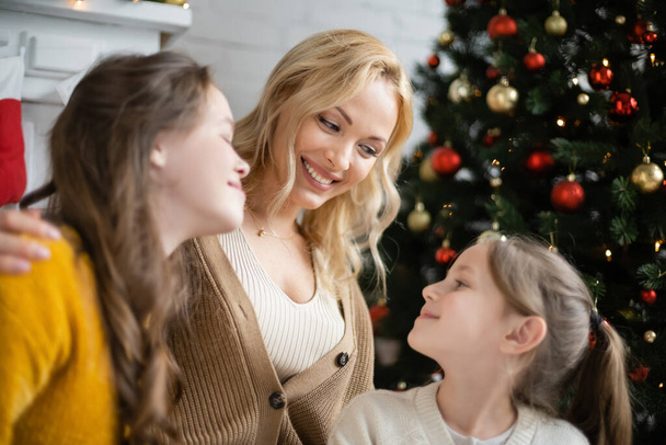 happy blonde woman looking at daughters near decorated christmas tree on blurred background - Foto, afbeelding