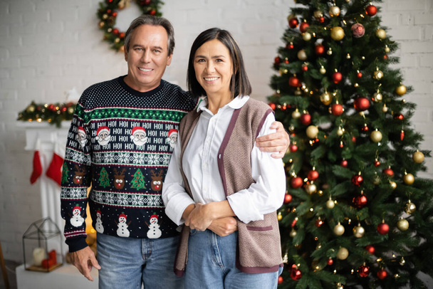 sonriente hombre en suéter con patrón festivo abrazando esposa multirracial cerca borrosa árbol de navidad - Foto, Imagen
