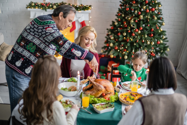 пожилой человек режет вкусную жареную индейку во время семейного ужина возле рождественской елки - Фото, изображение