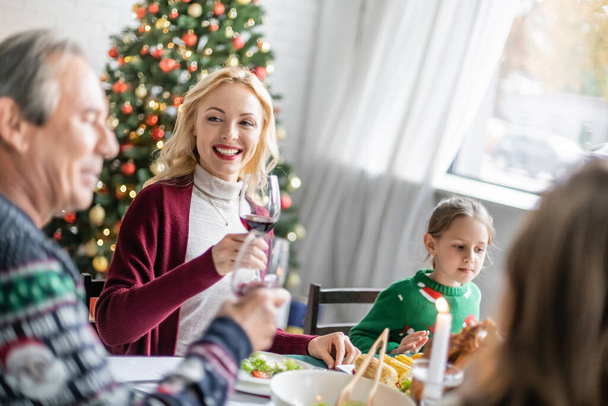 cheerful blonde woman toasting with glass of red wine near blurred father and daughters on christmas dinner - Photo, Image