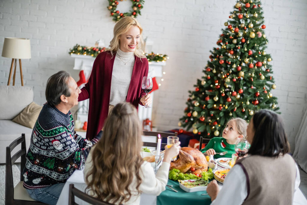 mujer sonriente tostada con copa de vino tinto durante la cena de Navidad con la familia interracial - Foto, imagen