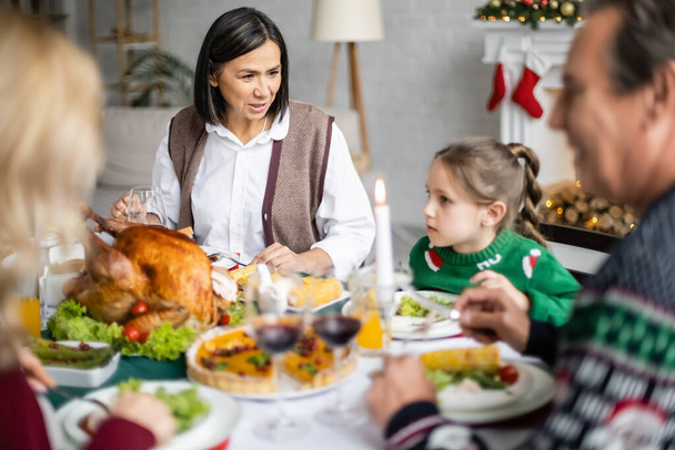 multiracial granny talking to granddaughter during christmas dinner with blurred family - Φωτογραφία, εικόνα