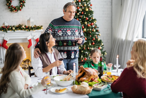 hombre mayor tostado con copa de vino tinto cerca de la familia multiétnica durante la cena de Navidad festiva - Foto, imagen