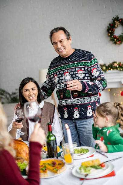 ευτυχισμένος ηλικιωμένος άνδρας που κάνει πρόποση με ένα ποτήρι κόκκινο κρασί ενώ γιορτάζει τα Χριστούγεννα με διαφυλετική οικογένεια - Φωτογραφία, εικόνα