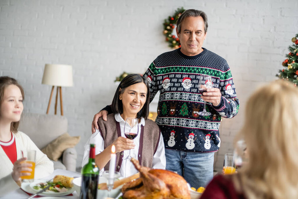 idősebb férfi és multiracionális nő pirítóssal, egy pohár vörösborral karácsonykor vacsora elmosódott családdal - Fotó, kép