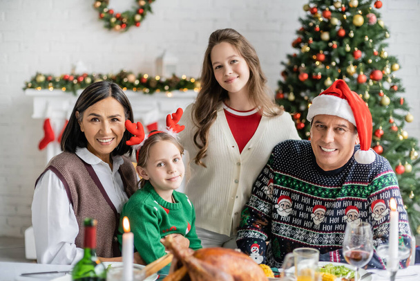 fröhliche Mädchen mit multiethnischen Großeltern, die in die Kamera schauen, in der Nähe von verschwommenem Festmahl und geschmücktem Weihnachtsbaum - Foto, Bild