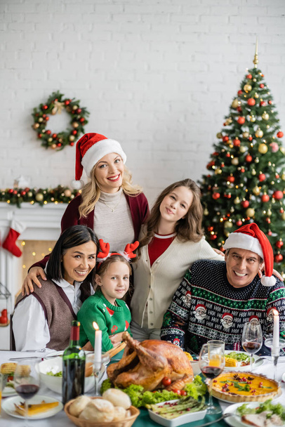 felice famiglia multietnica guardando la fotocamera vicino a cena festiva e albero di Natale a casa - Foto, immagini