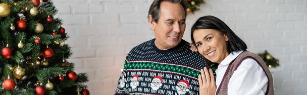 happy multiracial woman leaning on smiling husband near decorated christmas tree, banner - Foto, Imagen