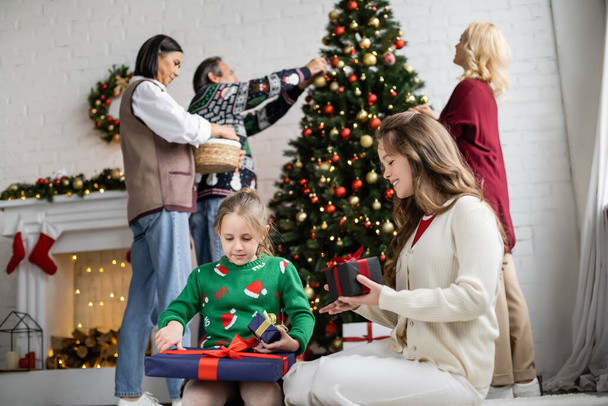 tytöt etsivät uutta vuotta lahjoja, kun taas rotujenvälinen perheen koristelu joulukuusi hämärtynyt tausta - Valokuva, kuva