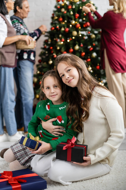 プレゼント箱の近くにお姉ちゃんを抱いてる10代の女の子背景にクリスマスツリーを飾る多民族家族 - 写真・画像