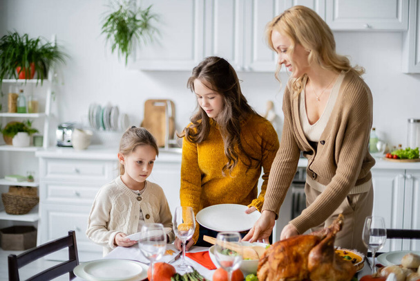 smiling blonde woman serving table for thanksgiving dinner with daughters in kitchen - Foto, Bild