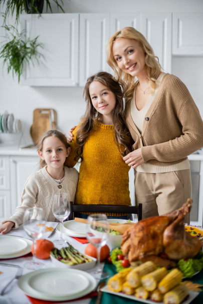 mother and daughters smiling at camera near table with delicious thanksgiving dinner - Fotó, kép