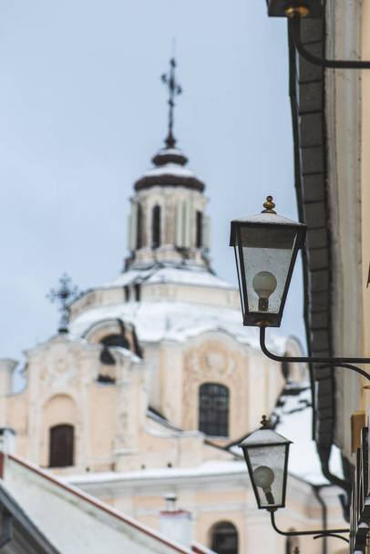 Hermosa calle estrecha en el casco antiguo de Vilna con la Iglesia Dominicana del Espíritu Santo, edificios antiguos y farolas en invierno cubierto por la nieve, vertical - Foto, Imagen