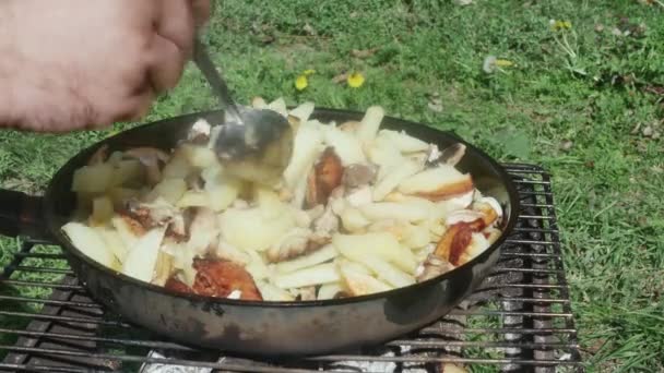  egy kéz keveri burgonya egy kéreg és a gomba egy serpenyőben egy grill, egy tisztáson egy kanállal. - Felvétel, videó