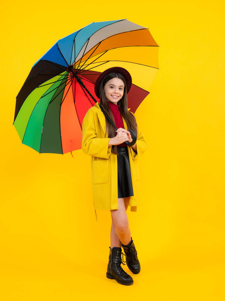 Child girl with rainbow umbrella in autumn weather isolated on yellow background. Autumn kids clothes. Smiling girl in autumn coat. Happy teenager portrait full length - Photo, Image
