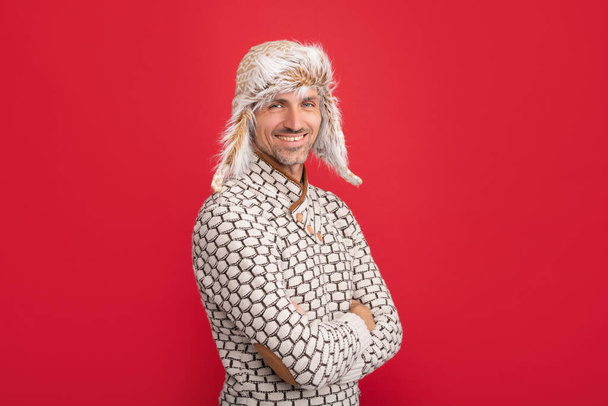 mannelijke winter mode. Mannen schoonheid. Een volwassen portret. Glimlachende man met oorflap hoed. grizzled man in trui op rode achtergrond. takelende man draagt warme hoed. - Foto, afbeelding