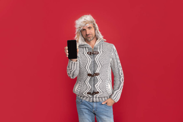 дорослий чоловік показує телефон. чоловіча модель моди тримає смартфон. концепція сучасного спілкування. грізний хлопець в светрі і капелюсі на червоному тлі
. - Фото, зображення