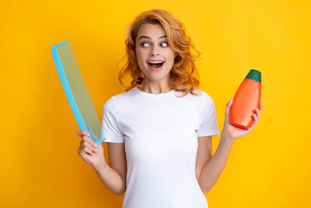 Surprised amazed girl combing hair. Redhead woman with a comb, isolated on yellow background. Woman hold bottle shampoo and conditioner - Photo, Image