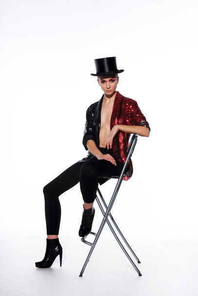 Full length image of a attractive young woman with makeup wear in a bright glitter costume and black top hat, asitting on high chair isolated white background. - Photo, Image